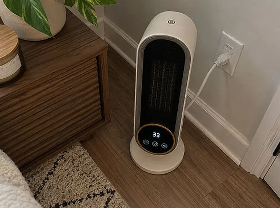 White Heatomi tower-style ceramic space heater with a round digital control panel, plugged into a wall outlet on a wood floor beside a nightstand
